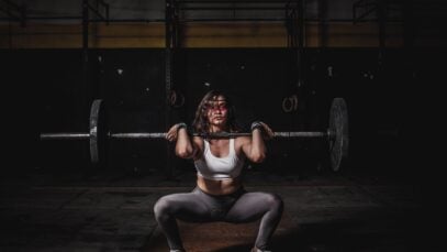 A strong female weightlifter squats with a barbell in a dimly lit gym, showcasing power and fitness.