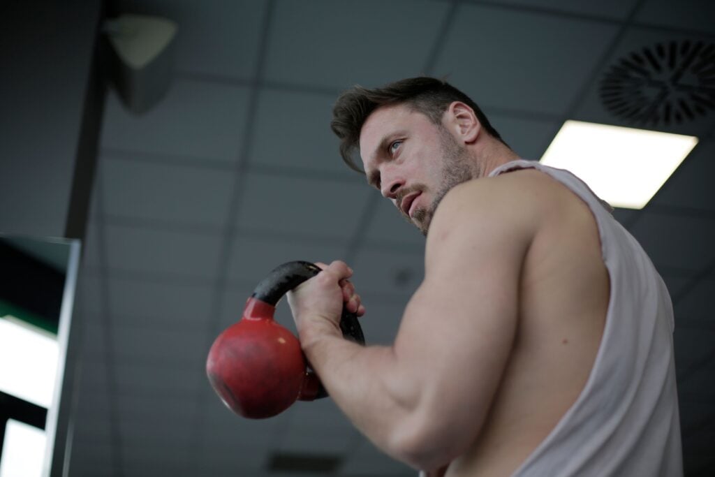 Low angle of adult male athlete wearing sportswear doing swing exercise with kettlebell in hand while training in sport center and looking over shoulder