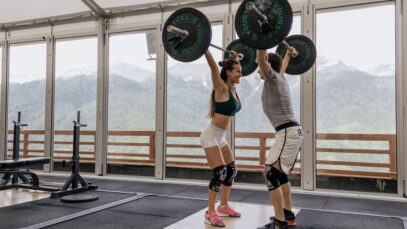 A couple lifting barbells together in a gym with scenic mountain views.