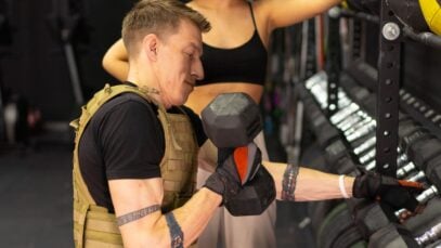 Side view of a man working out with a dumbbell and weighted vest in a gym setting.