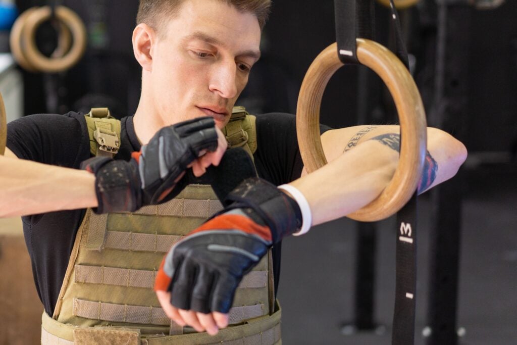 A man in tactical vest adjusting workout gloves next to gymnastic rings.